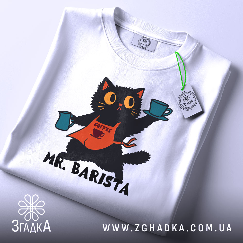 Футболка котик бариста, біла, складена на столі, фон світло-сірий, принт з котиком у фартуху, що тримає чашки з кавою, текст 'MR. BARISTA' під малюнком, на футболці є етикетка.