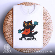 Футболка котик бариста, біла, складена на плетеній основі, фон світло-сірий, принт з котиком у фартуху, що тримає чашки з кавою, текст 'MR. BARISTA' під малюнком.