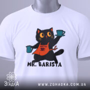 Футболка котик бариста, біла, складена на столі, фон світло-сірий, принт з котиком у фартуху, що тримає чашки з кавою, текст 'MR. BARISTA' під малюнком, на футболці є етикетка.