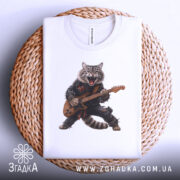 Футболка котик рокер гітарист, біла, складена на плетеній кошику, фон світло-сірий, принт з котом у рокерському стилі, який грає на гітарі, з енергійним виразом обличчя.