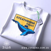 Футболка креативний відділ, біла, зображена в складеному вигляді, фон світло-сірий, принт з синім дельфіном у капелюсі та жовтим написом "КРЕАТИВНИЙ ВІДДІЛ", на футболці є етикетка з зеленим шнурком.