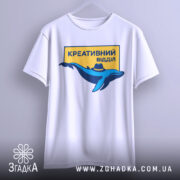 Футболка креативний відділ, біла, підвішена на вішалці, фон світло-сірий, принт з синім дельфіном у капелюсі та жовтим написом "КРЕАТИВНИЙ ВІДДІЛ".