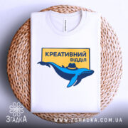 Футболка креативний відділ, біла, зображена в складеному вигляді на плетеній основі, фон світло-сірий, принт з синім дельфіном у капелюсі та жовтим написом "КРЕАТИВНИЙ ВІДДІЛ".