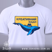 Футболка креативний відділ, біла, зображена в складеному вигляді, фон світло-сірий, принт з синім дельфіном у капелюсі та жовтим написом "КРЕАТИВНИЙ ВІДДІЛ", на футболці є етикетка з зеленим шнурком.