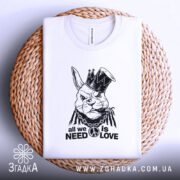 Футболка кролик панк, біла, зображена зверху на плетеній основі, на фоні світлого градієнта. Принт зображає кролика в короні з дредами, з написом 'all we need is love' під ним.