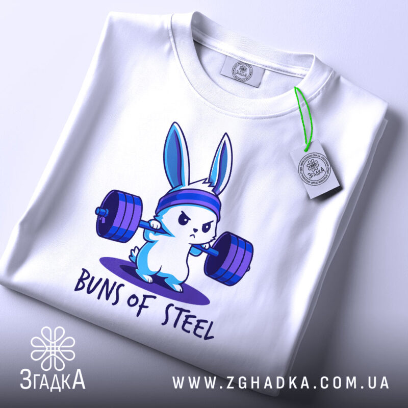 Футболка кролик сила, біла футболка, складена на столі, з принтом кролика, що піднімає штанги, на фоні світло-сірого кольору, з етикеткою зеленого кольору.