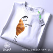 Футболка круасан гумор Glutton, біла, складена на столі, фон світлий, принт з чоловіком, що стоїть з круасаном на животі та написом 'GLUTTON', видима етикетка з зеленим шнурком.