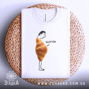 Футболка круасан гумор Glutton, біла, складена на плетеній кошику, фон світлий, принт з чоловіком, що стоїть з круасаном на животі та написом 'GLUTTON'.