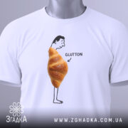 Футболка круасан гумор Glutton, біла, складена на столі, фон світлий, принт з чоловіком, що стоїть з круасаном на животі та написом 'GLUTTON', видима етикетка з зеленим шнурком.