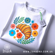 Футболка круасан у квітах, біла, зображена зверху, на столі, фон світло-сірий, принт з жовтим круасаном, кольоровими квітами та зеленим листям, на комірці є етикетка.