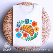 Футболка круасан у квітах, біла, складена на плетеній кошику, фон світло-сірий, принт з жовтим круасаном, кольоровими квітами та зеленим листям, видно комірець.