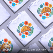 Футболка круасан у квітах, біла, зображена з фронтального ракурсу на манекені, фон світло-сірий, принт з жовтим круасаном оточеним кольоровими квітами та зеленим листям.