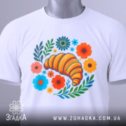 Футболка круасан у квітах, біла, зображена зверху, на столі, фон світло-сірий, принт з жовтим круасаном, кольоровими квітами та зеленим листям, на комірці є етикетка.