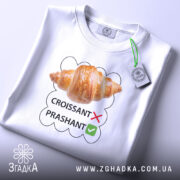 Футболка круасанний гумор Prashant, білого кольору, складена на столі, фон світло-сірий, принт з зображенням круасана та текстом 'CROISSANT' з червоним хрестиком і 'PRASHANT' з зеленим галочкою, на футболці є етикетка з зеленим шнурком.