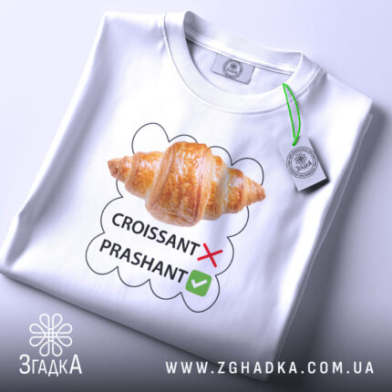 Футболка круасанний гумор Prashant, білого кольору, складена на столі, фон світло-сірий, принт з зображенням круасана та текстом 'CROISSANT' з червоним хрестиком і 'PRASHANT' з зеленим галочкою, на футболці є етикетка з зеленим шнурком.