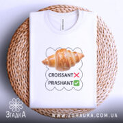 Футболка круасанний гумор Prashant, білого кольору, складена на плетеній кошику, фон світло-сірий, принт з зображенням круасана та текстом 'CROISSANT' з червоним хрестиком і 'PRASHANT' з зеленим галочкою.