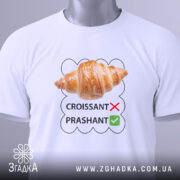 Футболка круасанний гумор Prashant, білого кольору, складена на столі, фон світло-сірий, принт з зображенням круасана та текстом 'CROISSANT' з червоним хрестиком і 'PRASHANT' з зеленим галочкою, на футболці є етикетка з зеленим шнурком.