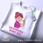 Футболка крихітка може все, біла футболка, складена на столі, з принтом дівчинки, яка показує м'язи, текст під малюнком фіолетового кольору, на футболці є етикетка з логотипом.