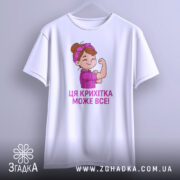 Футболка крихітка може все, біла футболка, підвішена на вішалці, з принтом дівчинки, яка показує м'язи, текст під малюнком написаний фіолетовим кольором, фон світло-сірий.