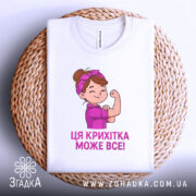 Футболка крихітка може все, біла футболка, складена на плетеному кошику, з принтом дівчинки, яка показує м'язи, текст під малюнком фіолетового кольору, фон світлий.