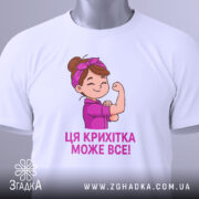 Футболка крихітка може все, біла футболка, складена на столі, з принтом дівчинки, яка показує м'язи, текст під малюнком фіолетового кольору, на футболці є етикетка з логотипом.