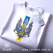 Футболка крила меч свободи, біла, складена на столі, фон світло-сірий, принт з мечем та крилами, на етикетці зелена стрічка.