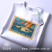 Футболка Кримський міст, біла, складена на столі, фон світло-сірий, принт у вигляді поштової марки з малюнком мосту, вантажівки та дронів, кольори принта: блакитний, оранжевий, жовтий, з етикеткою на шнурку.