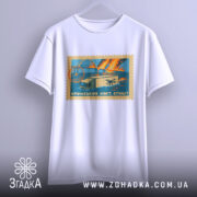 Футболка Кримський міст, біла, підвішена на вішалці, фон світло-сірий, принт у вигляді поштової марки з малюнком мосту, вантажівки та дронів, кольори принта: блакитний, оранжевий, жовтий.