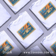 Футболка Кримський міст, біла, зображена на манекені, фон світло-сірий, принт у вигляді поштової марки з малюнком мосту, вантажівки та дронів, кольори принта: блакитний, оранжевий, жовтий.