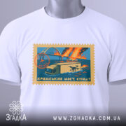 Футболка Кримський міст, біла, складена на столі, фон світло-сірий, принт у вигляді поштової марки з малюнком мосту, вантажівки та дронів, кольори принта: блакитний, оранжевий, жовтий, з етикеткою на шнурку.