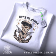 Футболка куля не дурна, біла, складена на поверхні, фон світло-сірий, принт з черепом у капелюсі, двома пістолетами та написом 'Куля не дурна', на футболці є етикетка з зеленим шнурком.