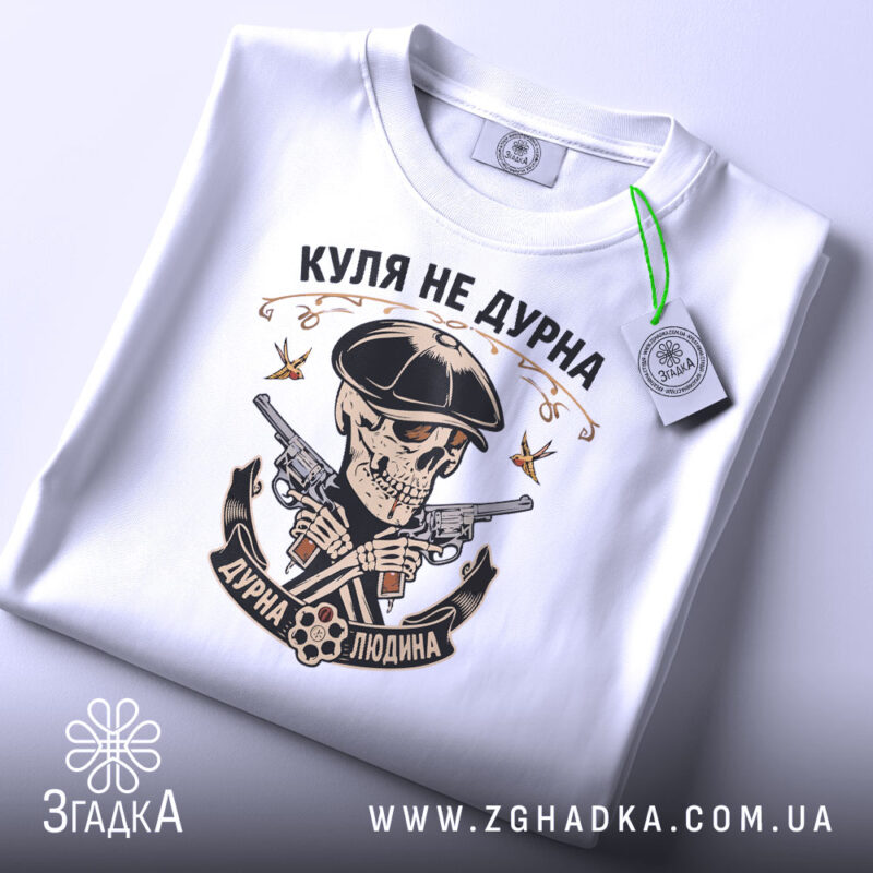 Футболка куля не дурна, біла, складена на поверхні, фон світло-сірий, принт з черепом у капелюсі, двома пістолетами та написом 'Куля не дурна', на футболці є етикетка з зеленим шнурком.