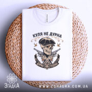 Футболка куля не дурна, біла, складена на плетеній кошику, фон світло-сірий, принт з черепом у капелюсі, двома пістолетами та написом 'Куля не дурна', видима етикетка на горловині.