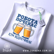 Футболка кум відпочиває, біла, складена на поверхні, фон світлий, принт з двома пивними кухлями та написом 'РОБОТА ПОЧЕКАЄ' у синьому кольорі, з зеленим ярликом.