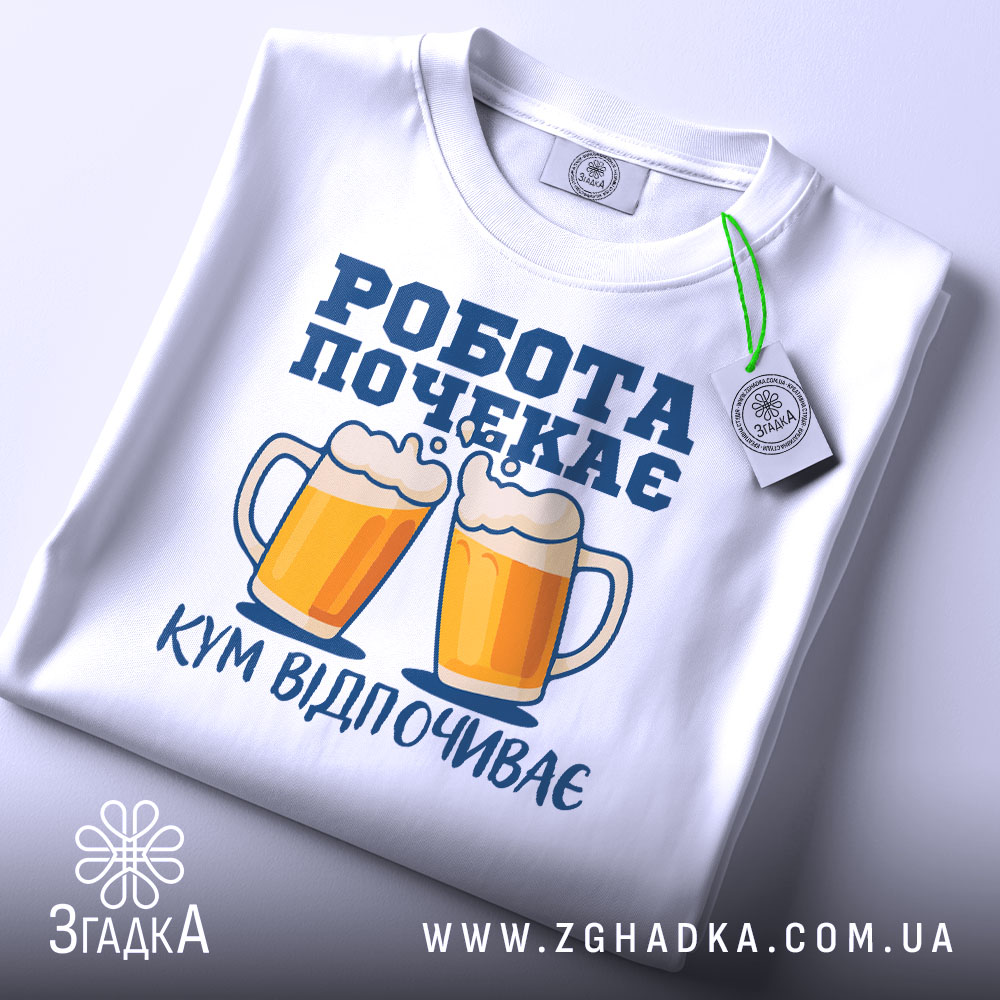 Футболка кум відпочиває, біла, складена на поверхні, фон світлий, принт з двома пивними кухлями та написом 'РОБОТА ПОЧЕКАЄ' у синьому кольорі, з зеленим ярликом.