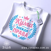 Футболка кума завжди права. Складена біла футболка, зображена зверху під кутом. На фоні світло-сірий градієнт. Принт у рожевих та блакитних кольорах, з написом 'Кума завжди права' та елементами декору, такими як корона та ваги.