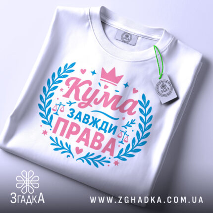Футболка кума завжди права. Складена біла футболка, зображена зверху під кутом. На фоні світло-сірий градієнт. Принт у рожевих та блакитних кольорах, з написом 'Кума завжди права' та елементами декору, такими як корона та ваги.