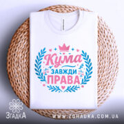 Футболка кума завжди права. Складена біла футболка, зображена зверху на плетеній кошику. На фоні світло-сірий градієнт. Принт у рожевих та блакитних кольорах, з написом 'Кума завжди права' та декоративними елементами, такими як корона та ваги.