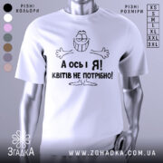 Футболка квітів не потрібно. Біла футболка з коротким рукавом, зображена на манекені з блискучою поверхнею. На футболці чорний принт з усміхненим персонажем і текстом "А ось і Я! Квітів не потрібно!". Фон світло-сірий, що підкреслює яскравість принта.