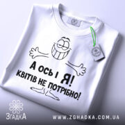 Футболка квітів не потрібно. Біла футболка, складена на плоскій поверхні, з видимим круглим вирізом. Принт з усміхненим персонажем і текстом "А ось і Я! Квітів не потрібно!" виконаний чорним кольором. На футболці також є етикетка з зеленим шнурком. Фон світло-сірий.