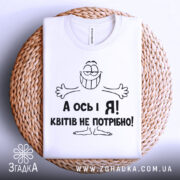 Футболка квітів не потрібно. Біла футболка, складена на плоскій поверхні, розташована на плетеній основі. Принт з усміхненим персонажем і текстом "А ось і Я! Квітів не потрібно!" виконаний чорним кольором. Фон світло-сірий, що підкреслює текстуру основи.