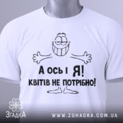 Футболка квітів не потрібно. Біла футболка, складена на плоскій поверхні, з видимим круглим вирізом. Принт з усміхненим персонажем і текстом "А ось і Я! Квітів не потрібно!" виконаний чорним кольором. На футболці також є етикетка з зеленим шнурком. Фон світло-сірий.