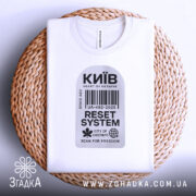 Футболка Київ білого кольору, складена на плетеній основі, фон світлий, принт у вигляді штрих-коду з написами 'Київ', 'RESET SYSTEM', 'CITY OF CHESTNUTS' та 'SCAN FOR FREEDOM', на комірці видна етикетка.
