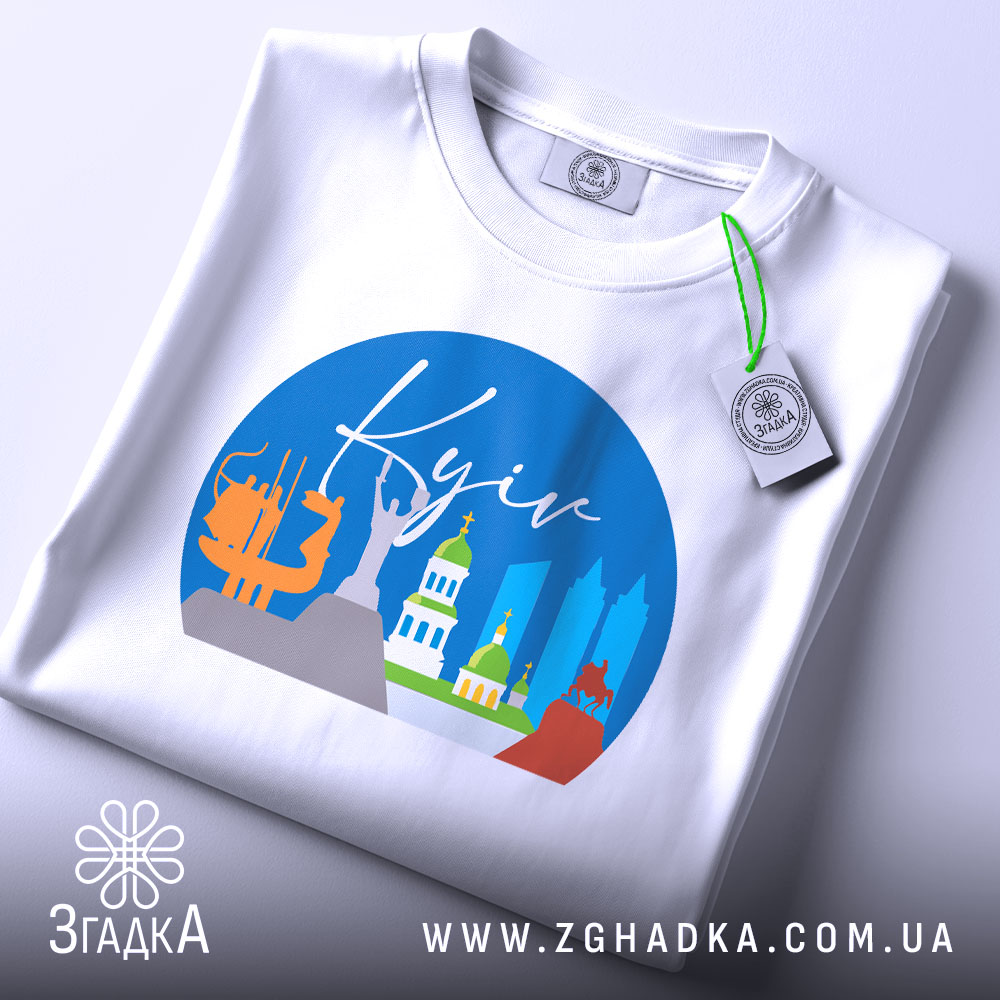 Футболка Київ міський стиль, біла, з верхнього ракурсу, складена на поверхні, фон світло-сірий, принт з зображенням архітектурних пам'яток Києва, з етикеткою на рукаві.