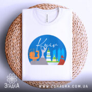 Футболка Київ міський стиль, біла, з верхнього ракурсу, складена на плетеній кошику, фон білий, принт з зображенням архітектурних пам'яток Києва в кольорах блакитного, жовтого, зеленого та червоного.