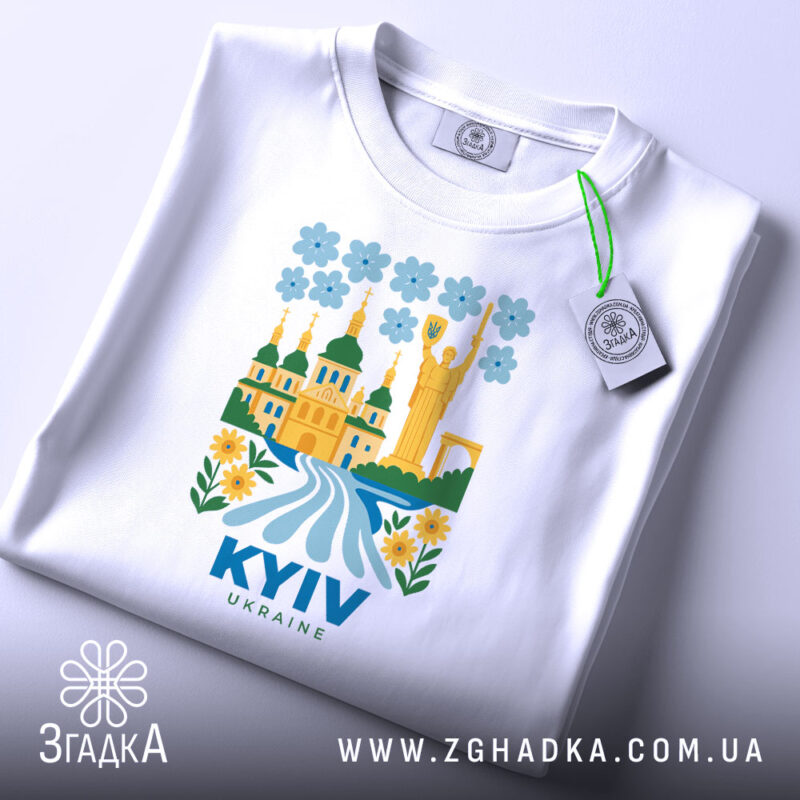 Футболка Київ білого кольору, складена на поверхні, з яскравим принтом, що зображує архітектуру Києва, квіти та річку, з етикеткою, що звисає з коміра, фон світло-блакитний.