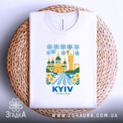 Футболка Київ білого кольору, складена на плетеній кошику, з яскравим принтом, що зображує архітектуру Києва, квіти та річку, фон світло-блакитний.