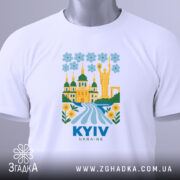 Футболка Київ білого кольору, складена на поверхні, з яскравим принтом, що зображує архітектуру Києва, квіти та річку, з етикеткою, що звисає з коміра, фон світло-блакитний.