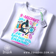 Футболка лаборант білого кольору, складена на столі, фон світло-сірий, принт з мікроскопом, пробірками та текстом, прикріплений етикеткою з зеленим шнурком.