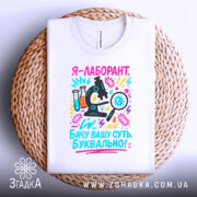 Футболка лаборант білого кольору, складена на плетеній основі, фон світло-сірий, принт містить мікроскоп, пробірки та текст, що підкреслює наукову тематику.